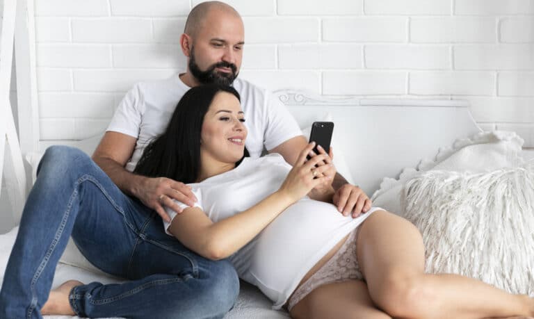 Mężczyzna obejmuje uśmiechniętą kobietę w ciąży, która trzyma telefon